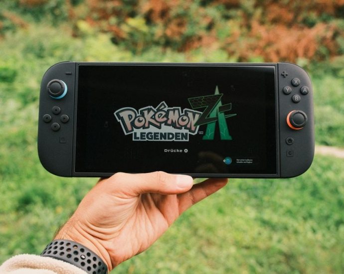 A hand holding a handheld gaming console displaying the Pokémon Legends game screen outdoors.